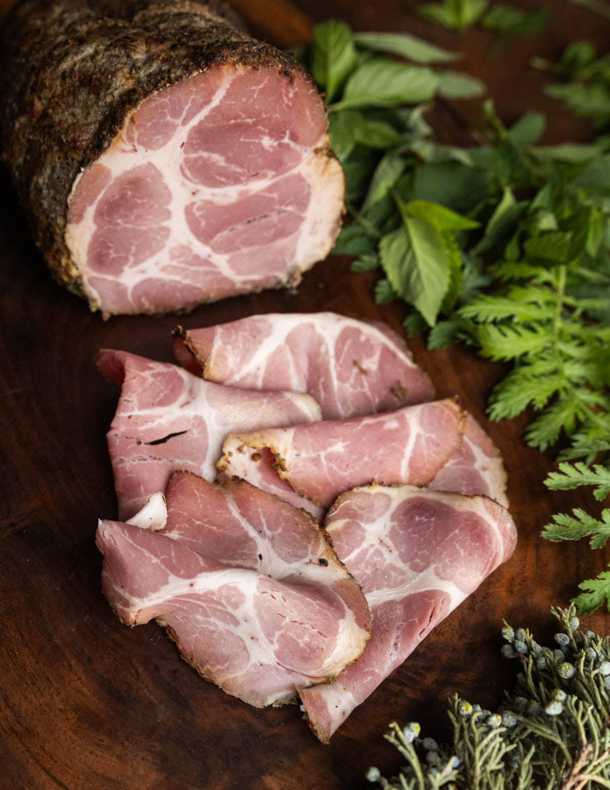 Sliced ham cured with wild herbs on a cutting board surrounded by yarrow, bee balm and wild juniper.