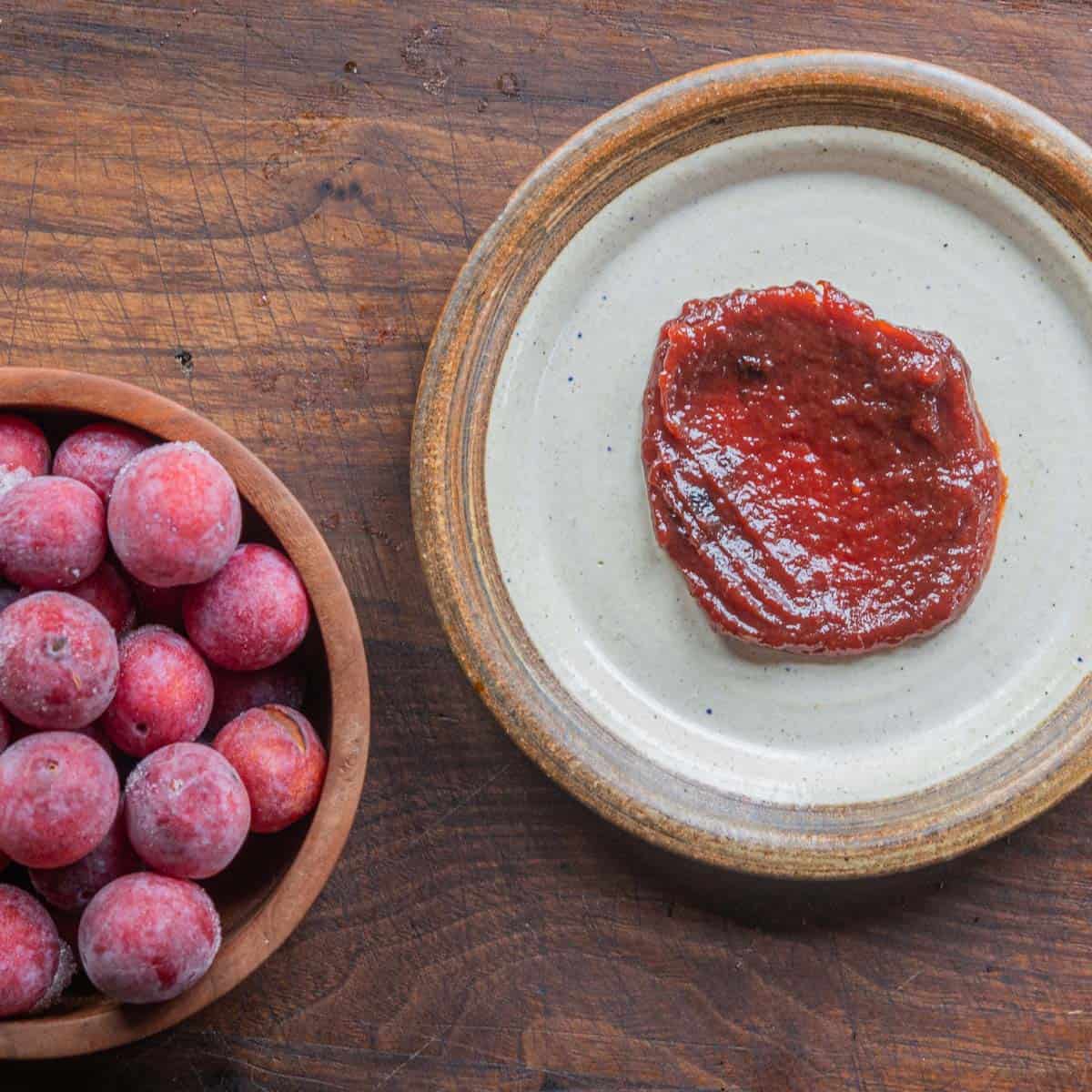 A jar of wild plum butter next to a plate of wild plum butter and a bowl of wild plums. 