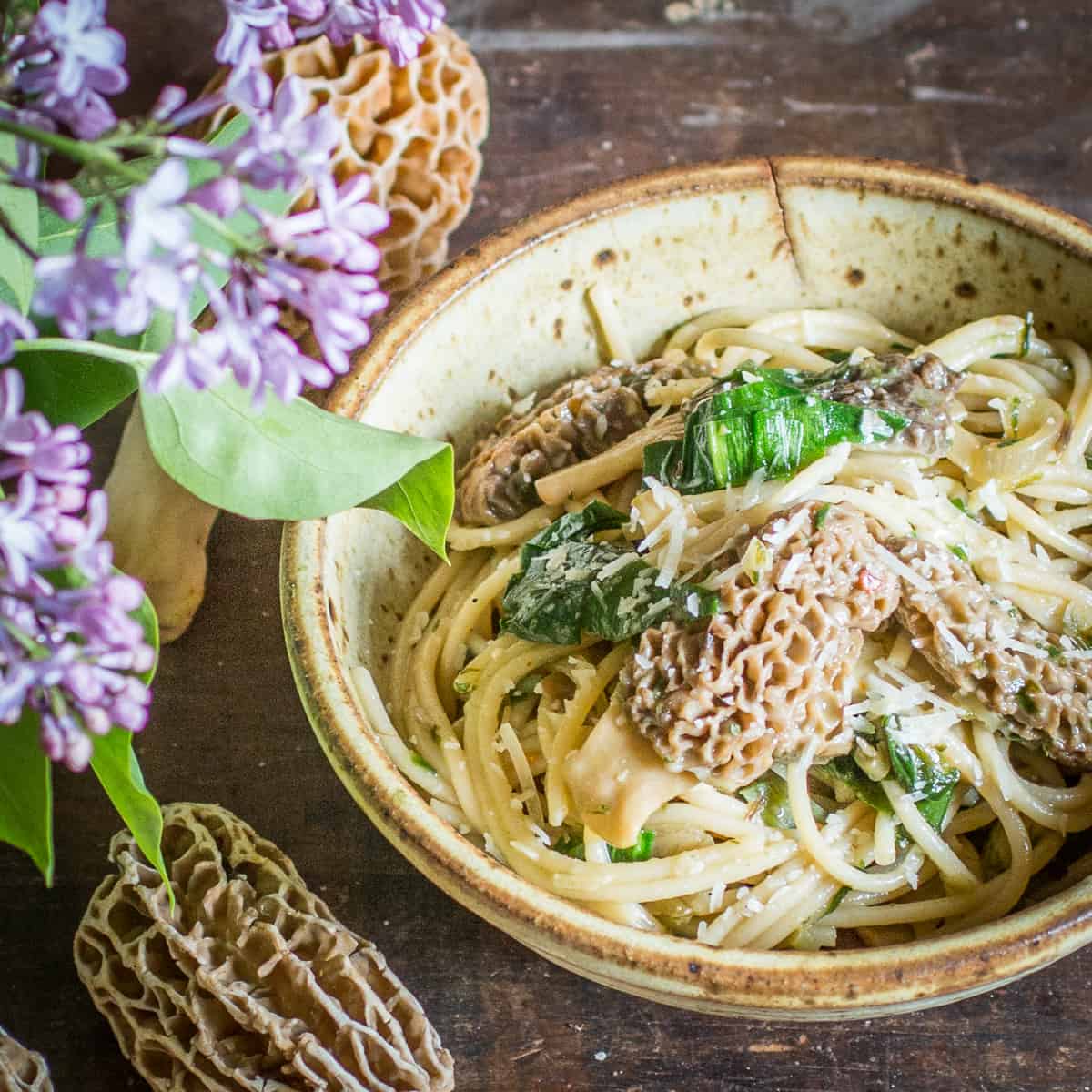 A bowl of morel mushroom pasta next to fresh morels and lilac flowers. 