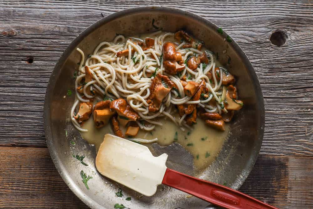 chanterelle mushroom spaghetti in an all clad pan on grey wood background with red spatula 