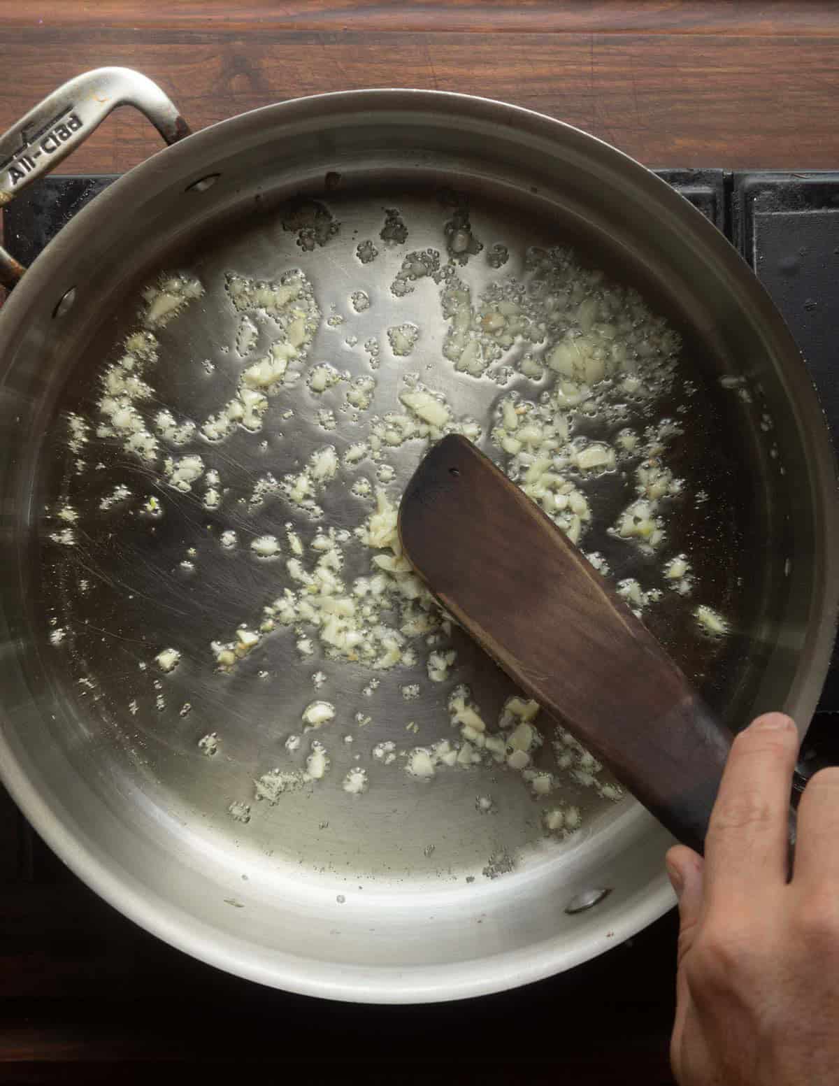 Sweating minced garlic in a large all clad pan stirring with a wooden spatula.
