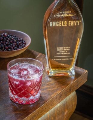 A glass of chokecherry cocktail next to a bottle of bourbon and a bowl of wild cherries.