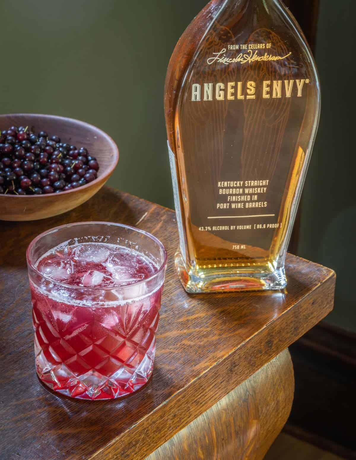 A glass of chokecherry cocktail next to a bottle of bourbon and a bowl of wild cherries. 