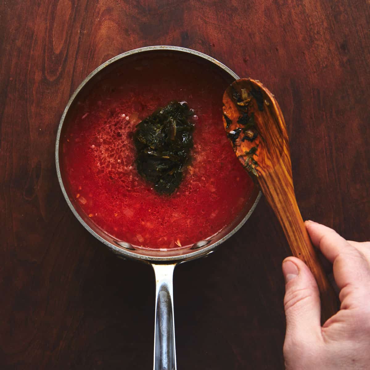 Adding dried greens to a pot of soup with a wooden spoon.