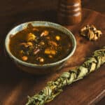 A bowl of soup next to a braid of dried greens, a peppermill, and a few nuts.