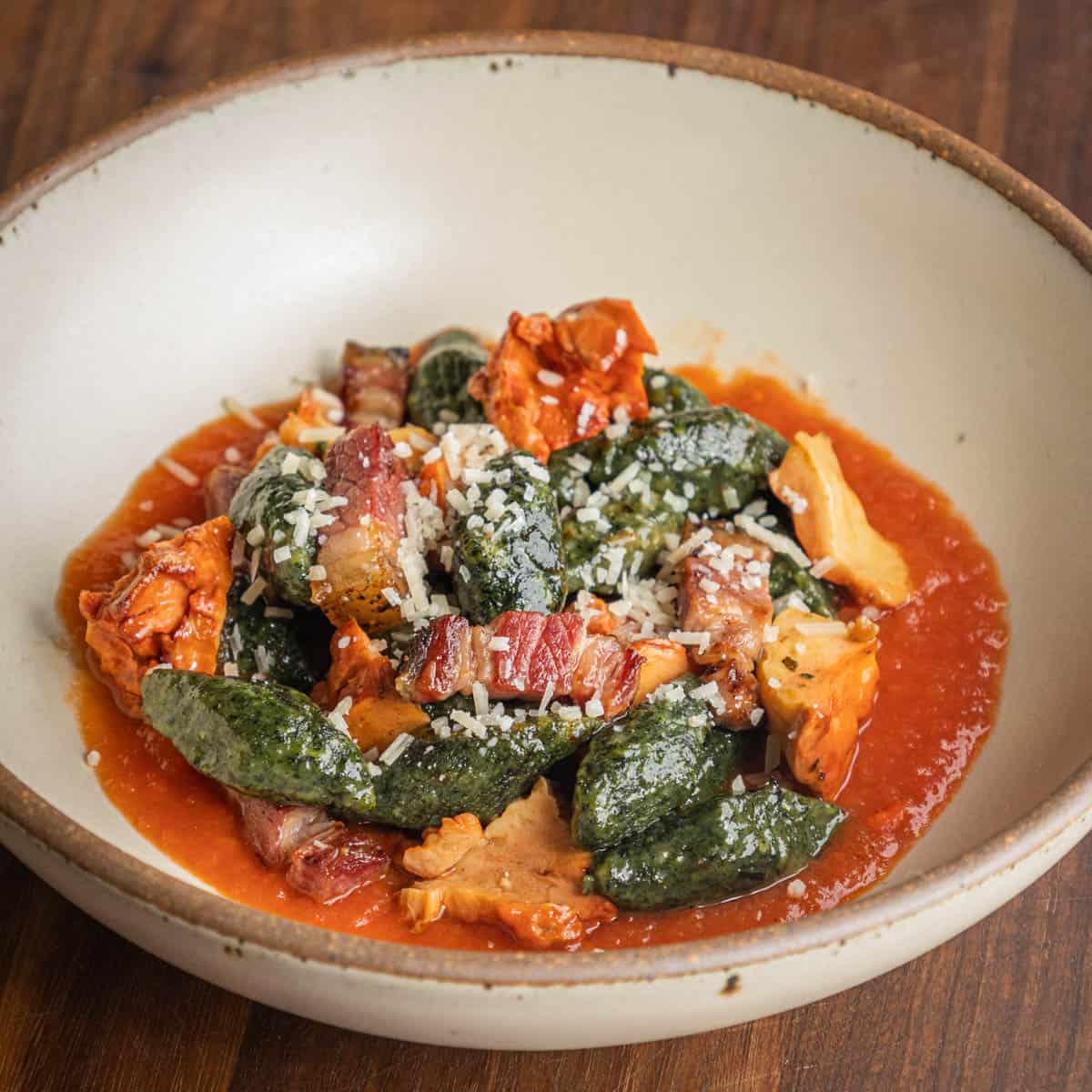 A bowl of green gnocchi with beef bacon and mushrooms on a bed of tomato sauce.