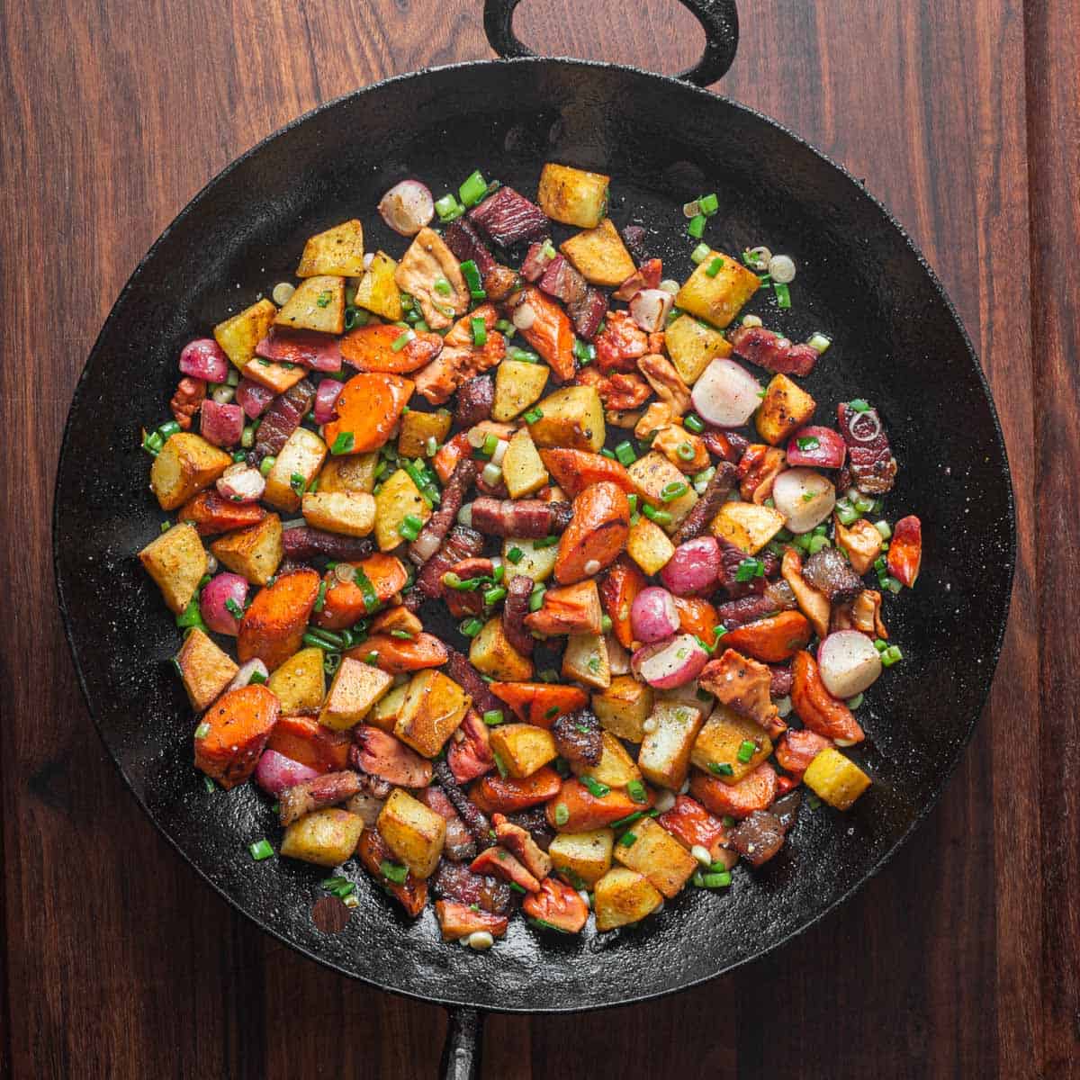 A carbon steel pan full of root vegetable and potato hash with beef bacon and mushrooms.