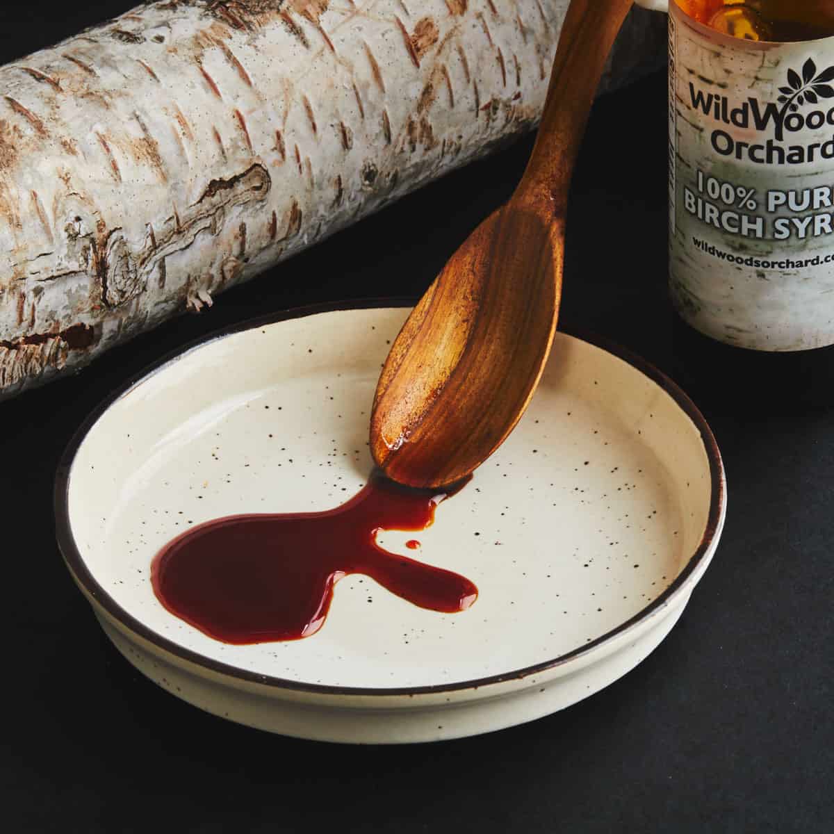 A white plate with birch syrup drizzling off a spoon next to a birch log.