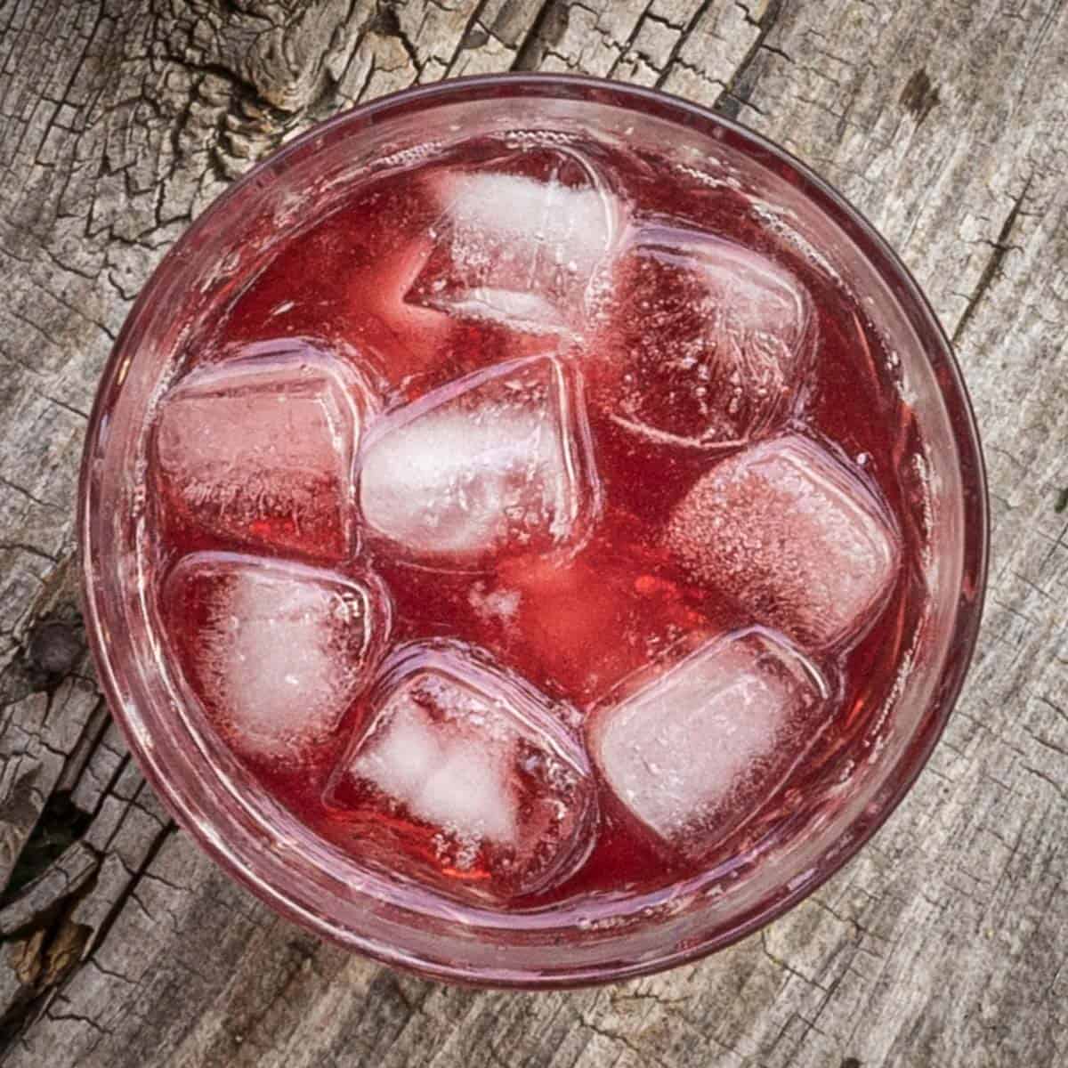 A glass filled with a red cocktail and ice on a grey wood background.