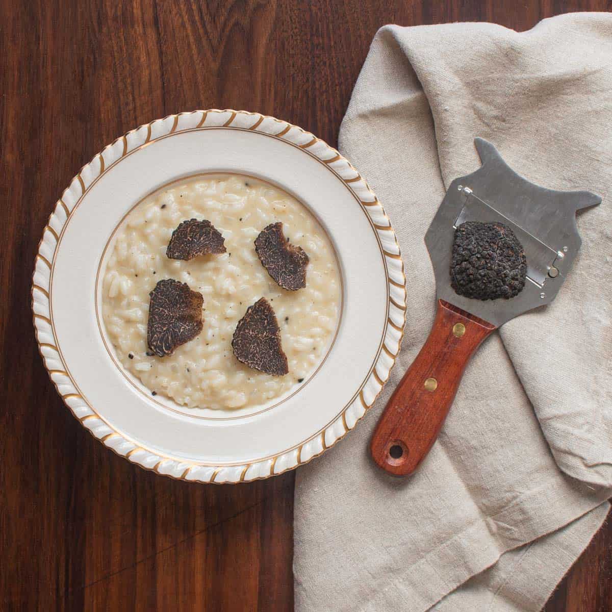 White bowl of black truffle risotto next to a napkin and truffle slicer.