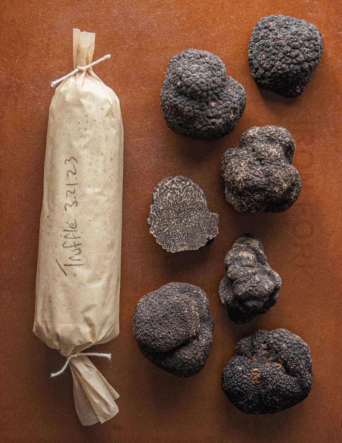 A round log of black truffle butter rolled in parchment next to many black truffles. 