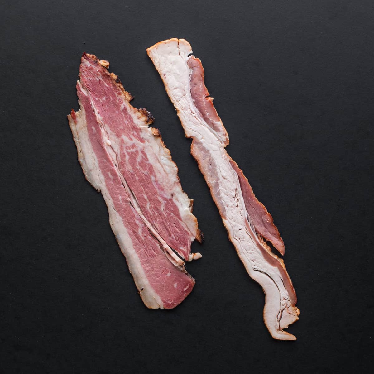 A slice of raw beef bacon next to pork bacon on a black background.