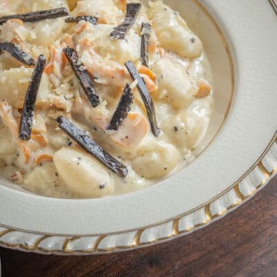 A close up image of a bowl of gnocchi with mushrooms in a creamy sauce topped with fresh sliced black truffles next to a napkin.