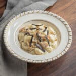 A bowl of gnocchi with mushrooms in a creamy sauce topped with fresh sliced black truffles next to a napkin.