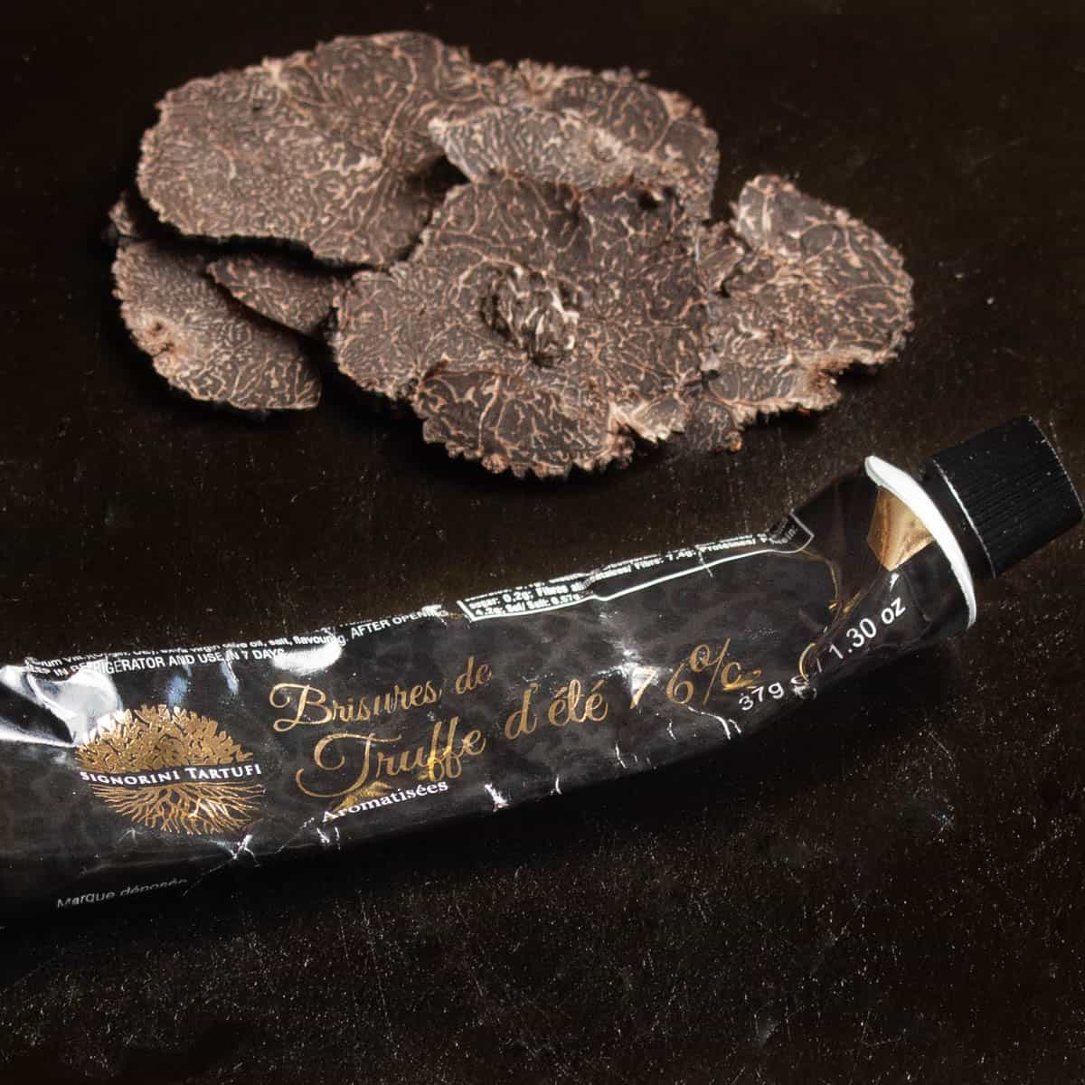 A tube of truffle paste or pate on a black board next to shaved black truffles. 