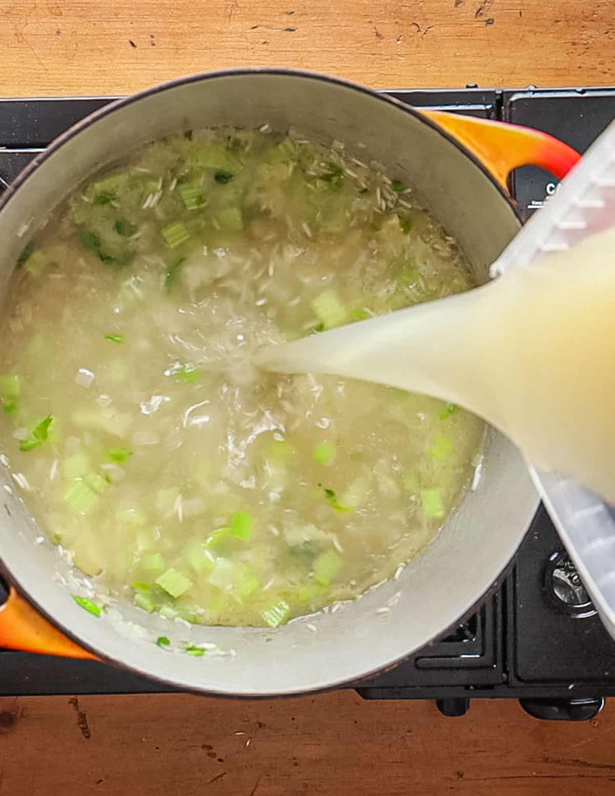 Pouring chicken stock into a pot of cooking rice, shallots and celery.
