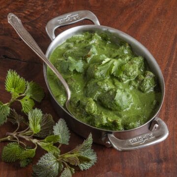 A green lamb saag curry in a dish with a silver spoon on a wooden background surrounded by soi, or nettles.