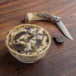 A bowl of creamy sausage rigatoni covered with pecorino cheese and shaved black truffles next to a truffle and a truffle knife.