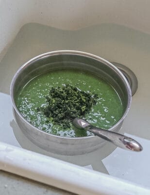 Chilling stinging nettle soup in a metal bowl placed in a sink of cold water and adding chopped nettle greens.