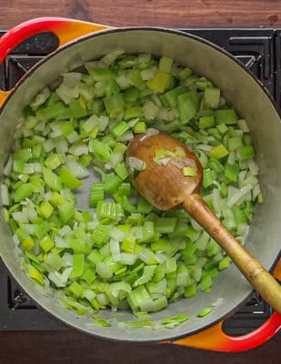 A top-down image of cooking celery, onion and leek with butter using a wooden spoon in a soup pot.