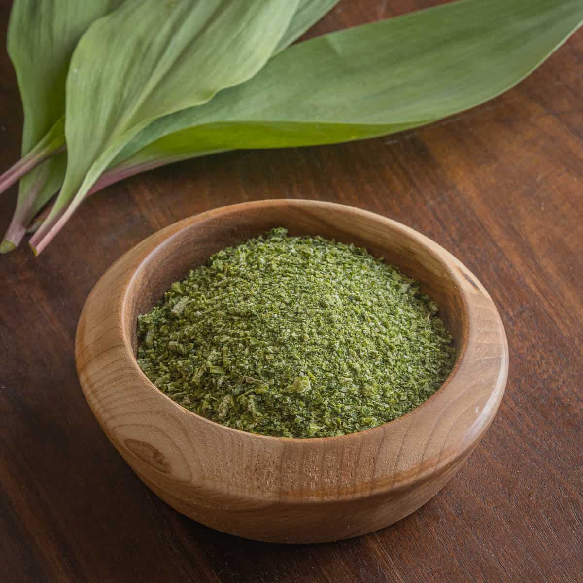 a bowl of green ramp salt next to fresh ramp leaves on a wooden board.