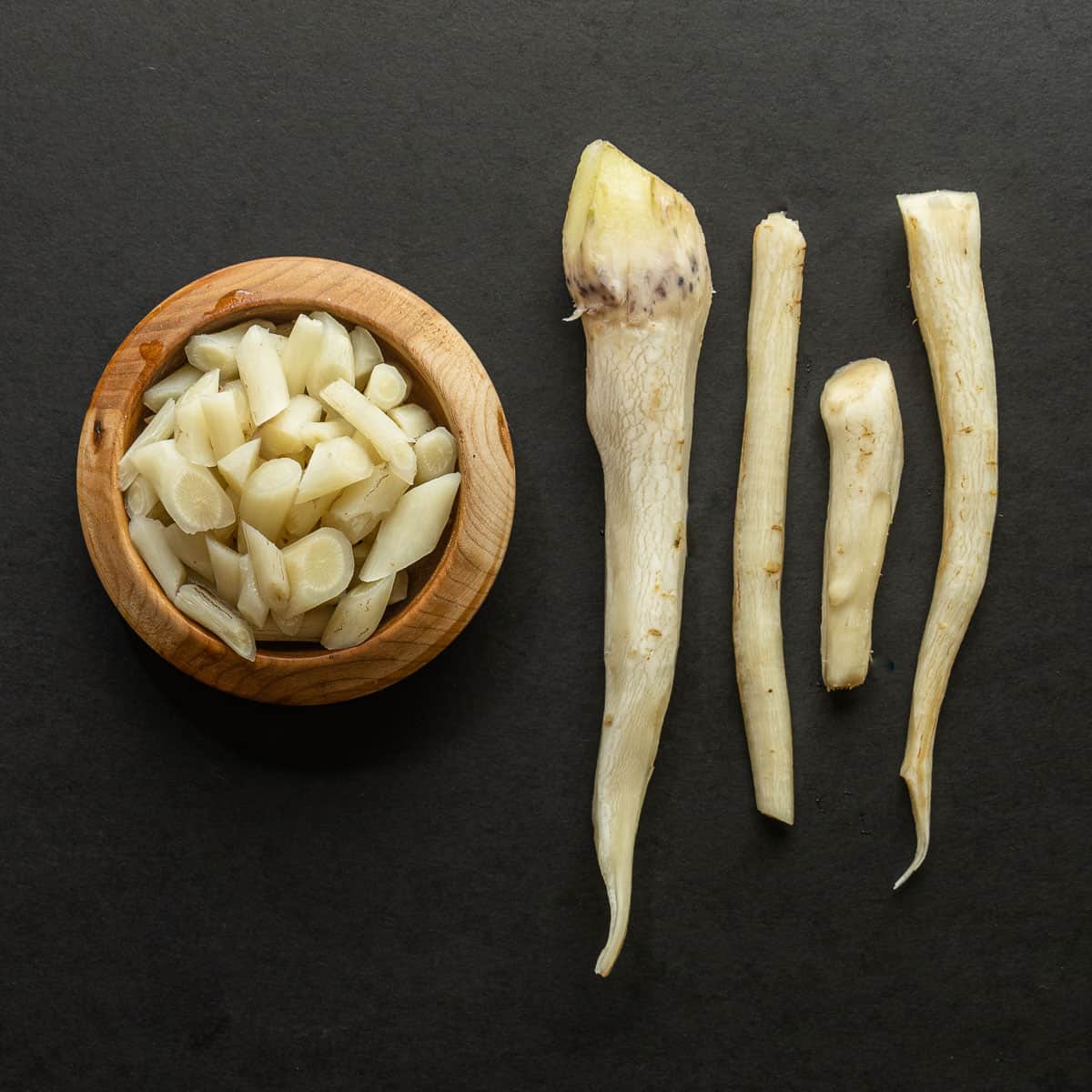 A bowl of cooked thistle roots next to peeled thistle roots on a black background. 