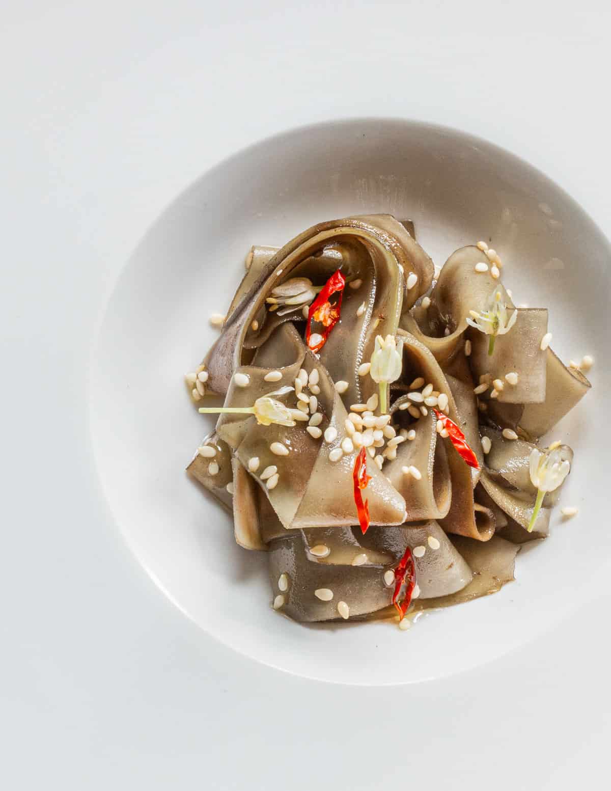 A bowl of brown burdock noodles garnished with chili and ramp flowers. 
