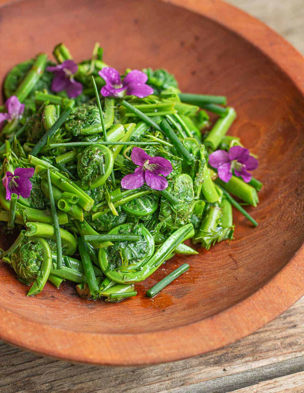 A wooden bowl filled with a salad of wild fiddlehead ferns garnished with wild violet flowers. 