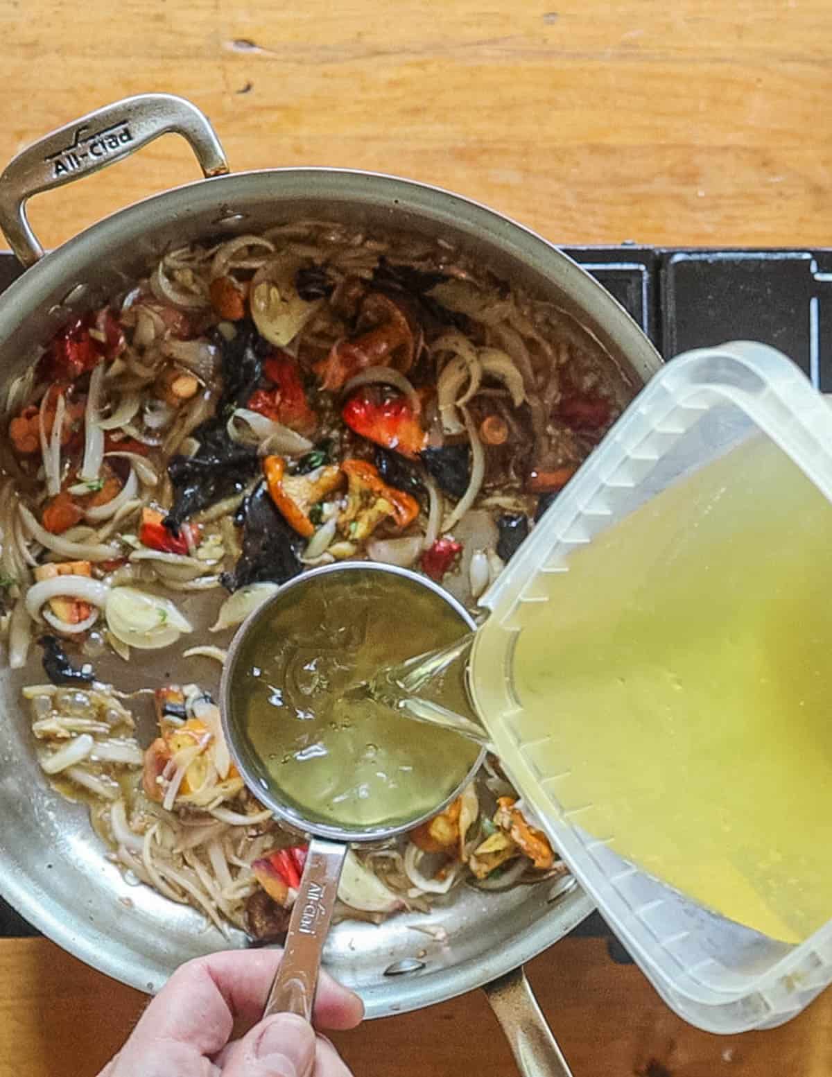 Pouring chicken stock into a pan of cooked mushrooms and shallots. 