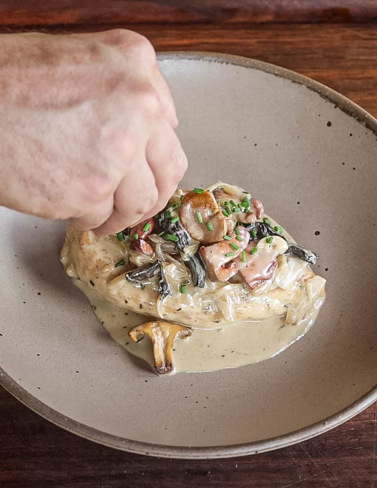 Sprinkling chives on top of a cooked chicken breast with mushroom sauce. 