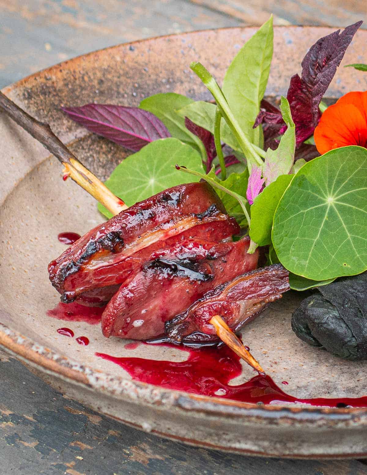 Pigeon meat and bacon skewers on a plate drizzled with wild cherry sauce next to a salad. 