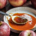 A spoon filled with apple cider molasses on a plate surrounded by apples.