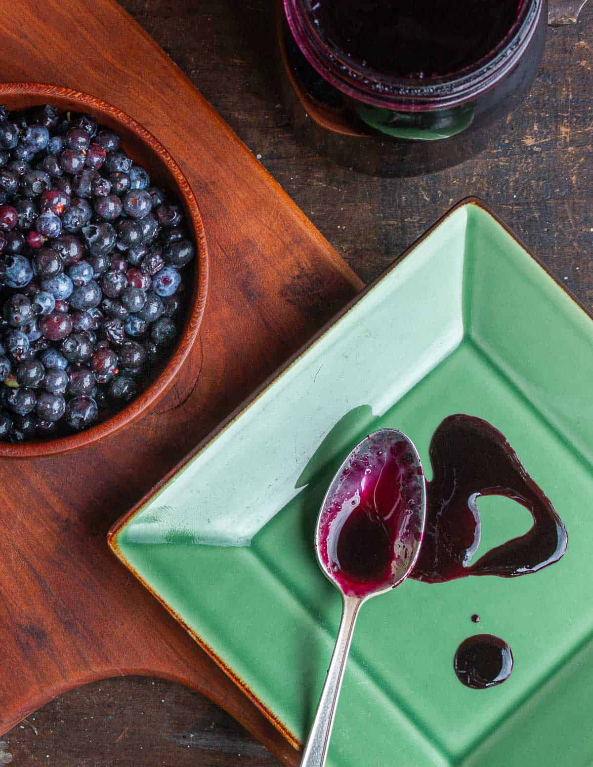 Wild blueberry syrup on a plate next to a bowl of blueberries and a jar of blueberry molasses. 