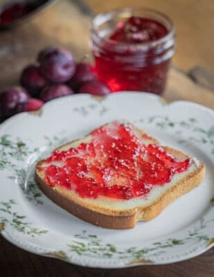 A plate with a piece of toast with crab apple jelly with crab apples and a jar of jelly in the background. 
