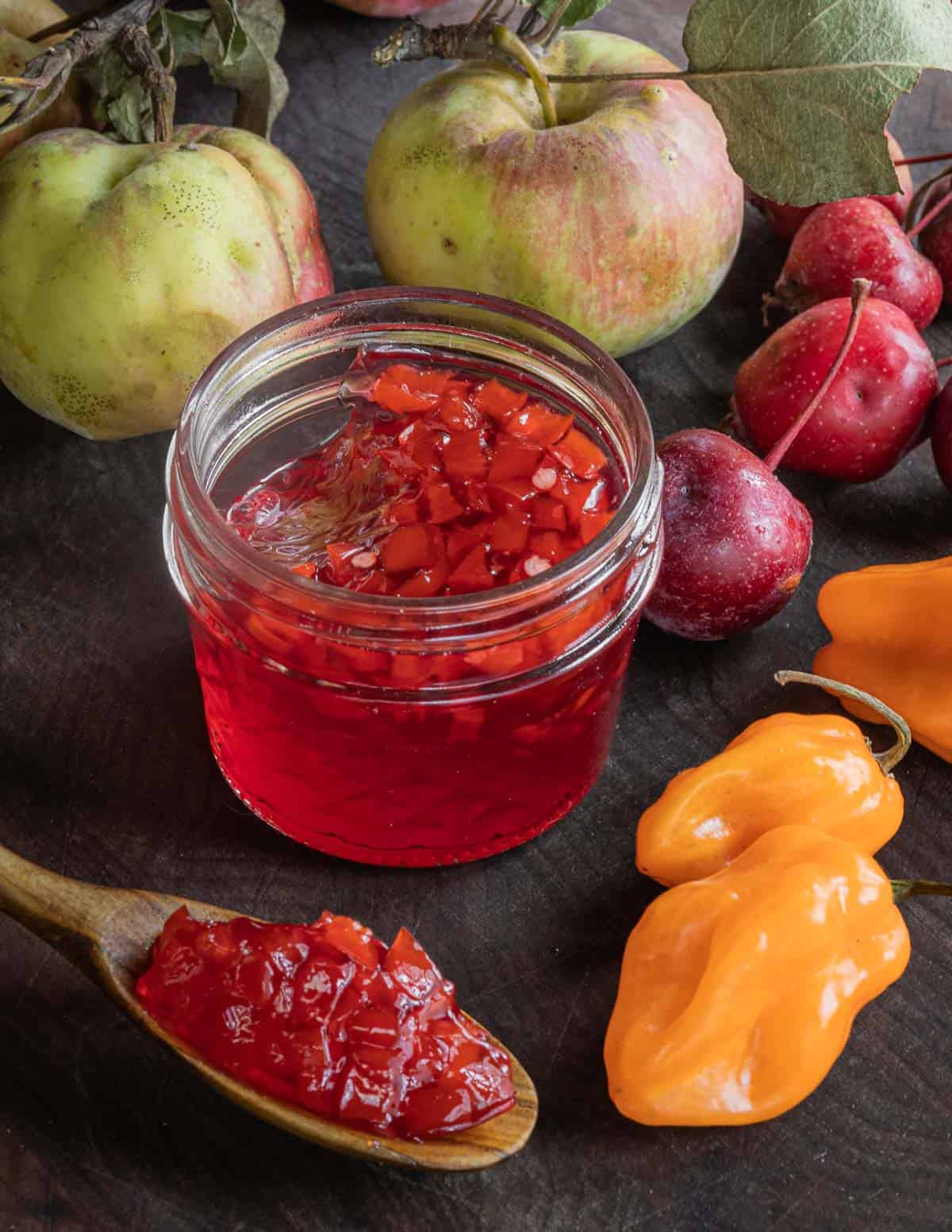 A jar of crab apple hot pepper jelly made with habaneros next to fresh peppers and apples. 