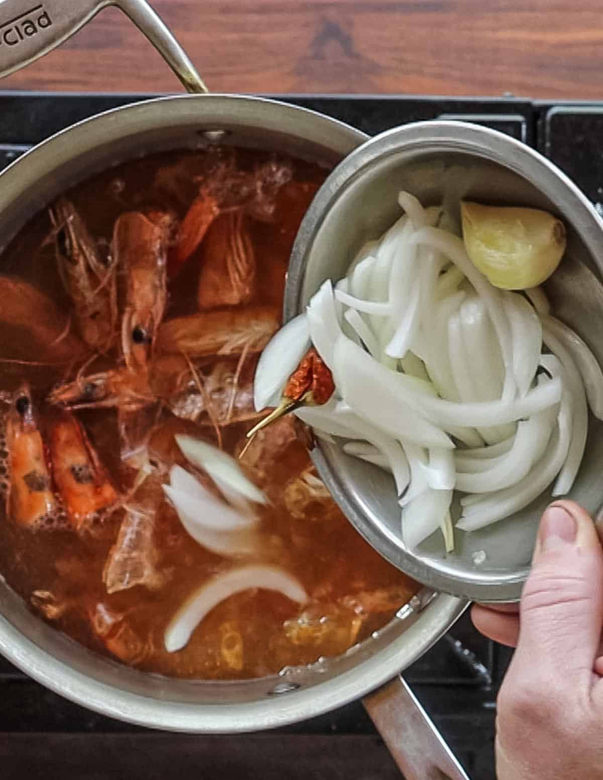 Adding sliced onion, galangal, lemongrass and garlic to a pot of shrimp heads, shells and water. 
