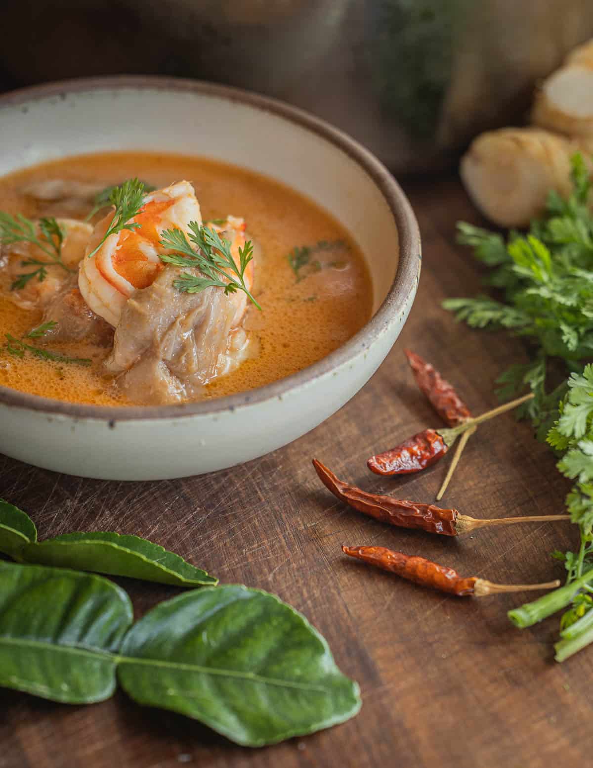 A bowl of Thai mushroom soup with shrimp and coconut milk in a bowl surrounded by Thai chilis, Kaffir lime leaves cilantro, lime and galangal. 