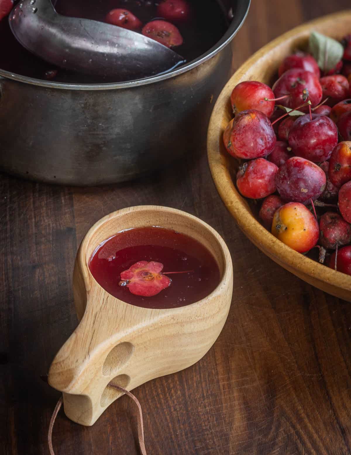 A mug filled with red crab apple cider next to a bowl of crab apples and a pot of crab apple cider. 