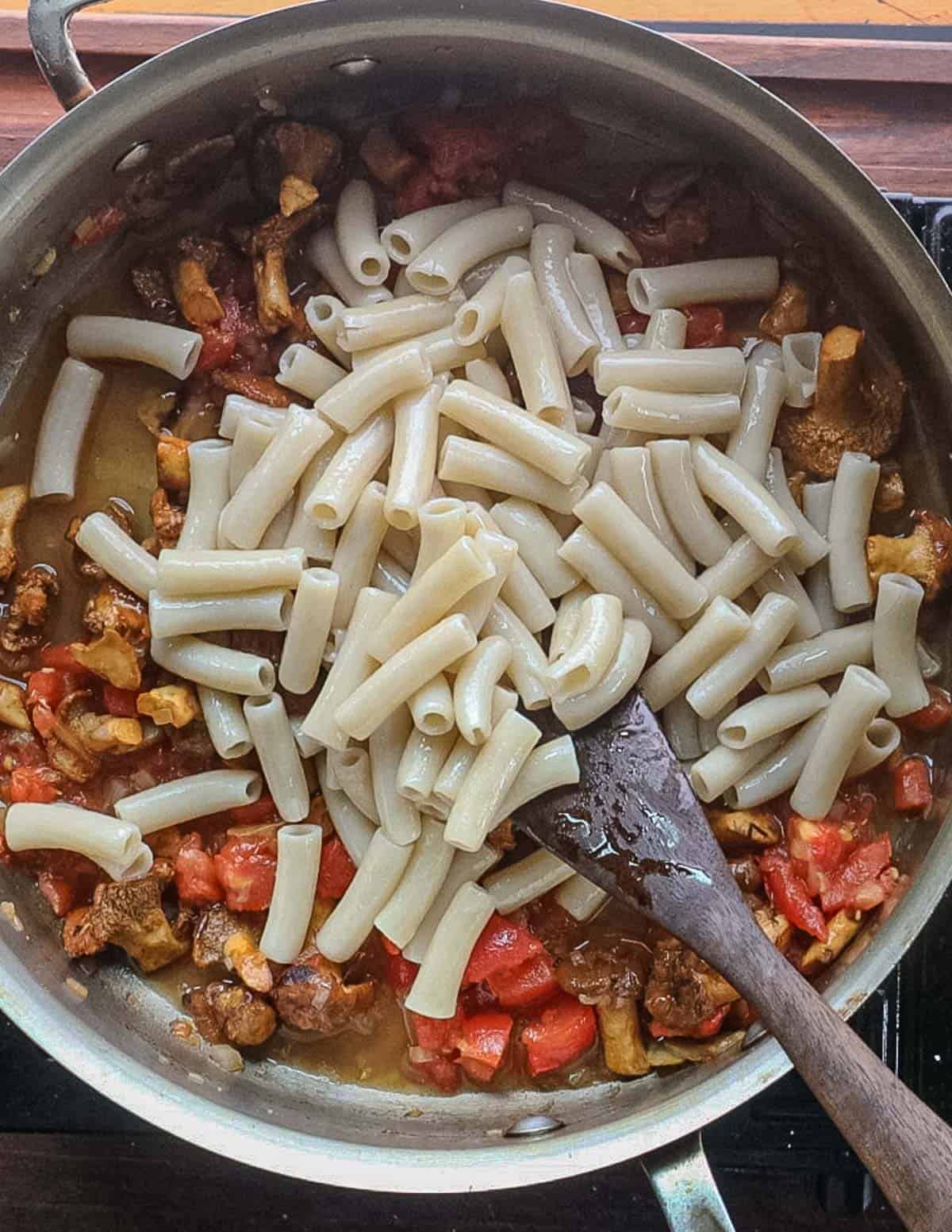 Adding cooked ziti pasta to a pan of mushrooms, tomato, shallots and garlic. 