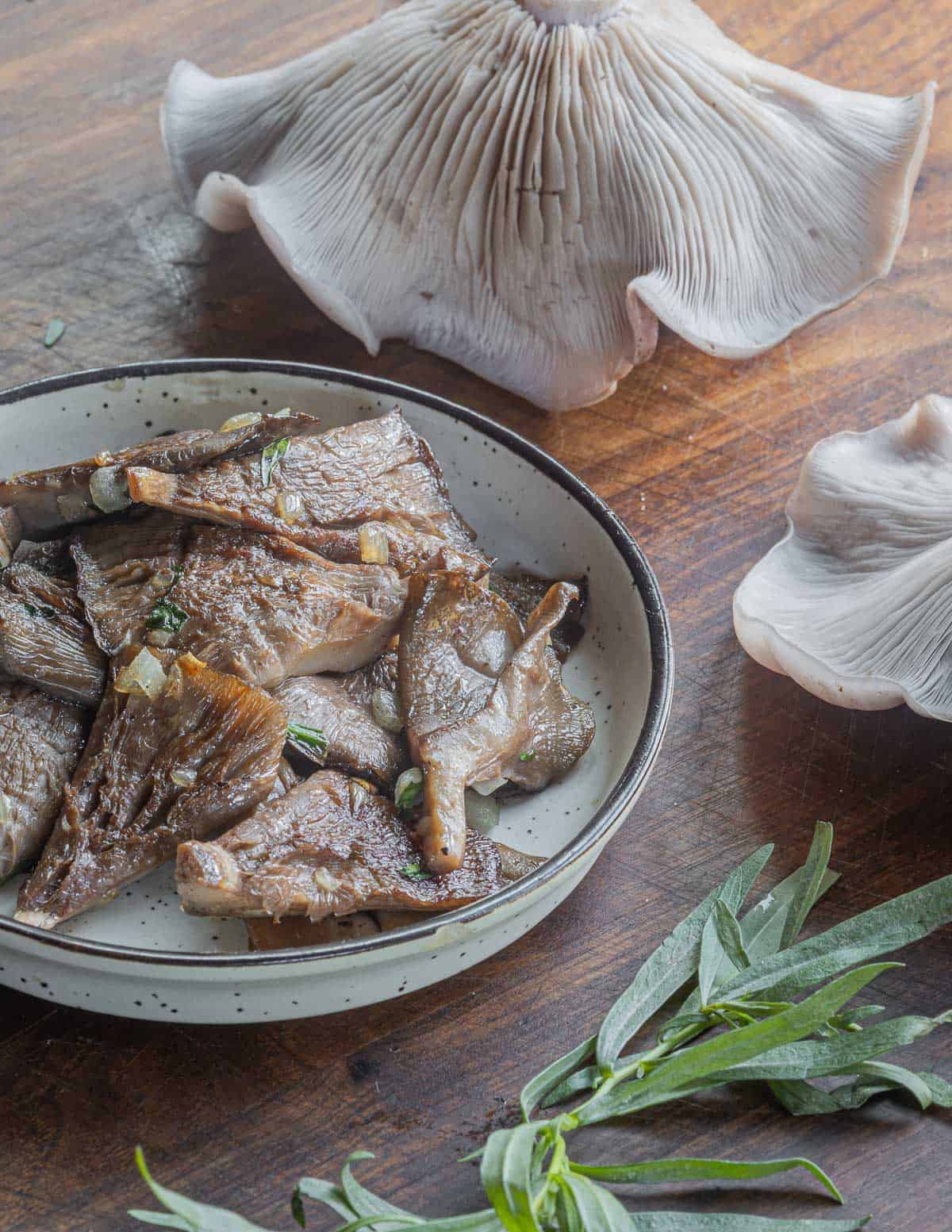 A small bowl of sauteed mushrooms next to fresh wood blewit mushrooms and a sprig of tarragon. 