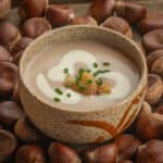 A bowl of Italian chestnut soup garnished with creme fraiche, chopped cooked chestnuts and chives surrounded by fresh chestnuts.