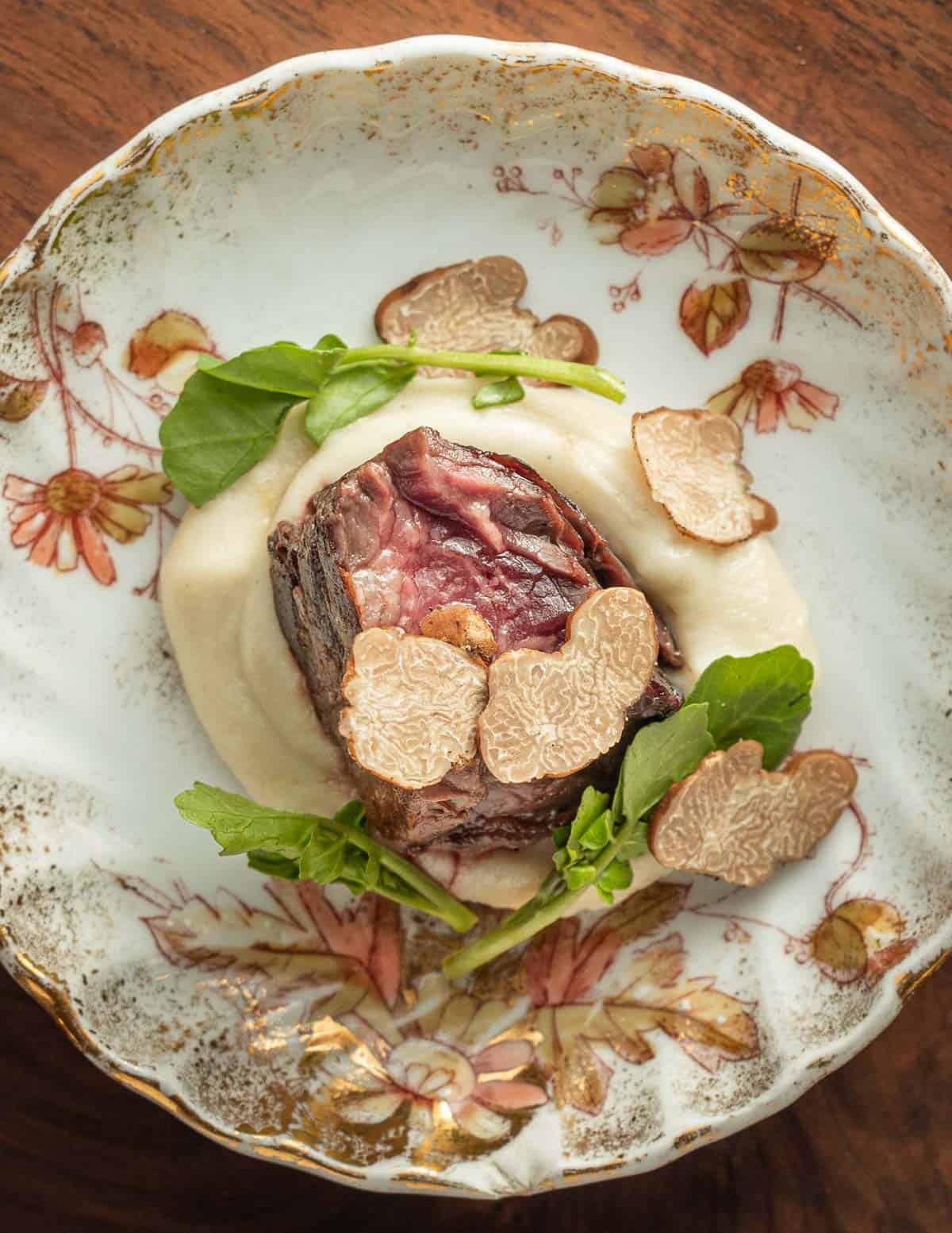 A cooked piece of hanger steak on a China plate next to sunchoke puree, watercress, and fresh sliced pecan truffles.