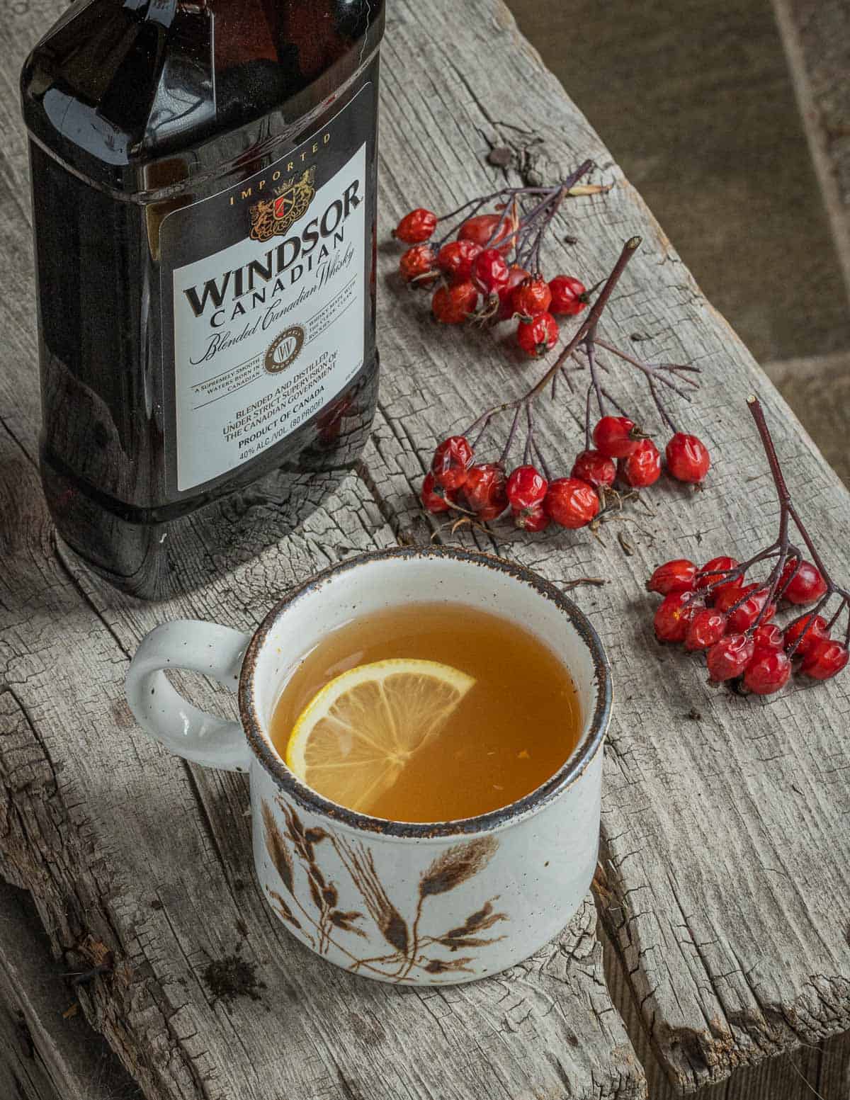A rosehip hot toddy in a ceramic mug with wheat on it next to a bottle of windsor whiskey and fresh clusters of wild rose hips. 