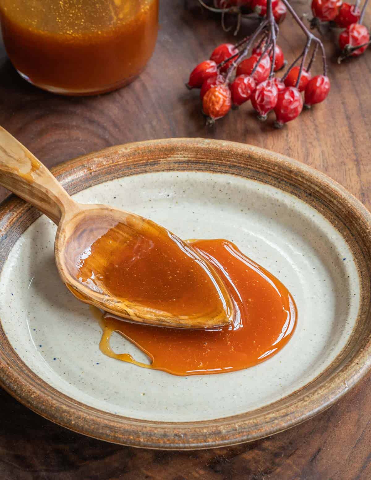 An applewood spoon with rosehip syrup on a plate next to fresh rosehips and jar of syrup.