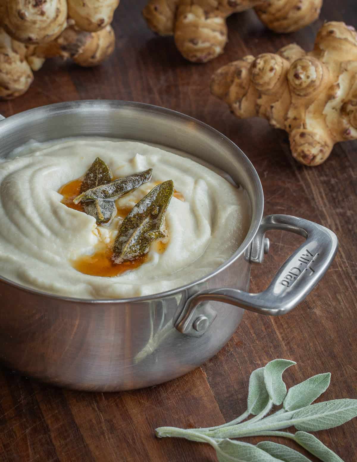 A small pan of white sunchoke puree surrounded by raw sunchokes, sage, and fried sage leaves in brown butter. 