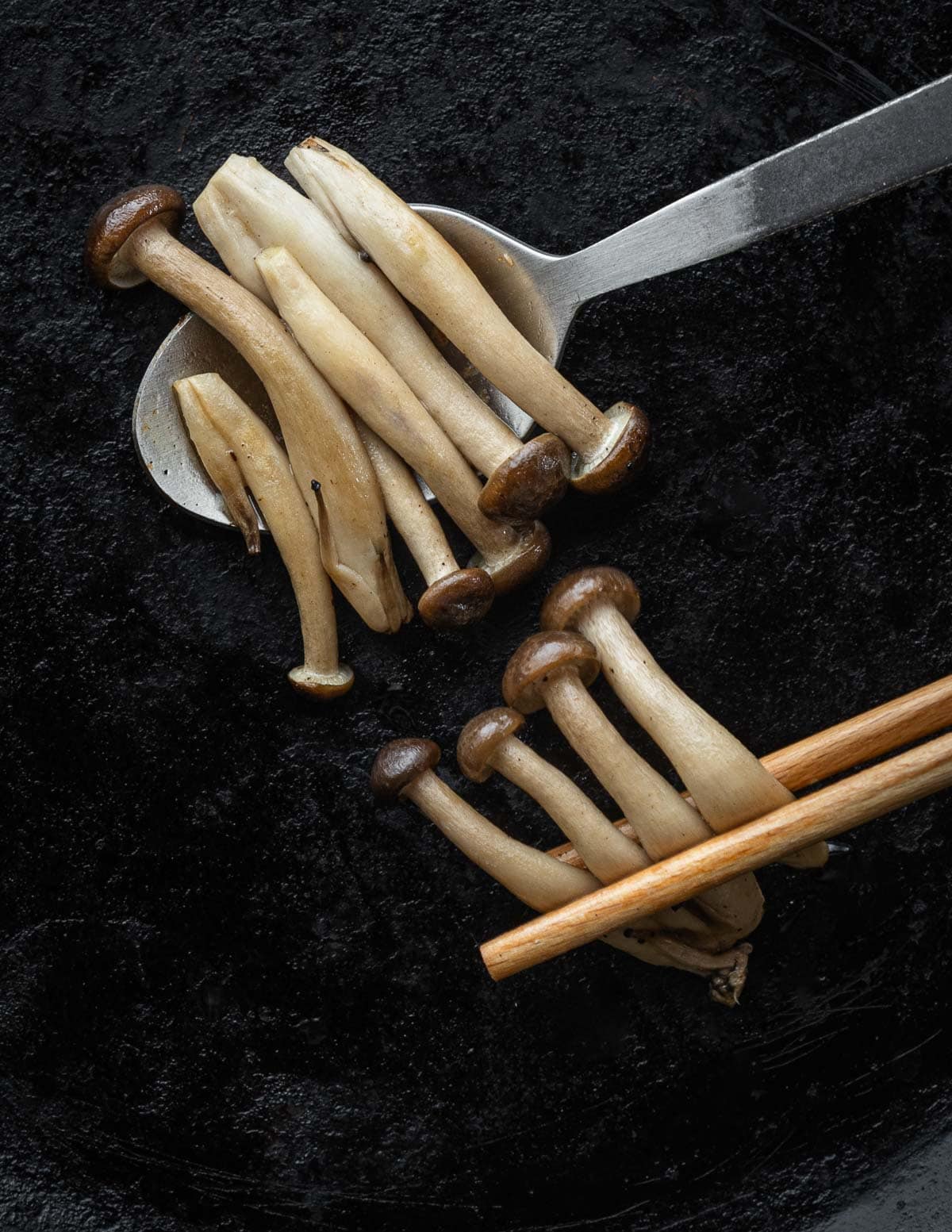 Cooked beech mushrooms falling off of a spoon on a black background, shown next to mushrooms neatly held by chopsticks to illustrate traditional cooking methods. 