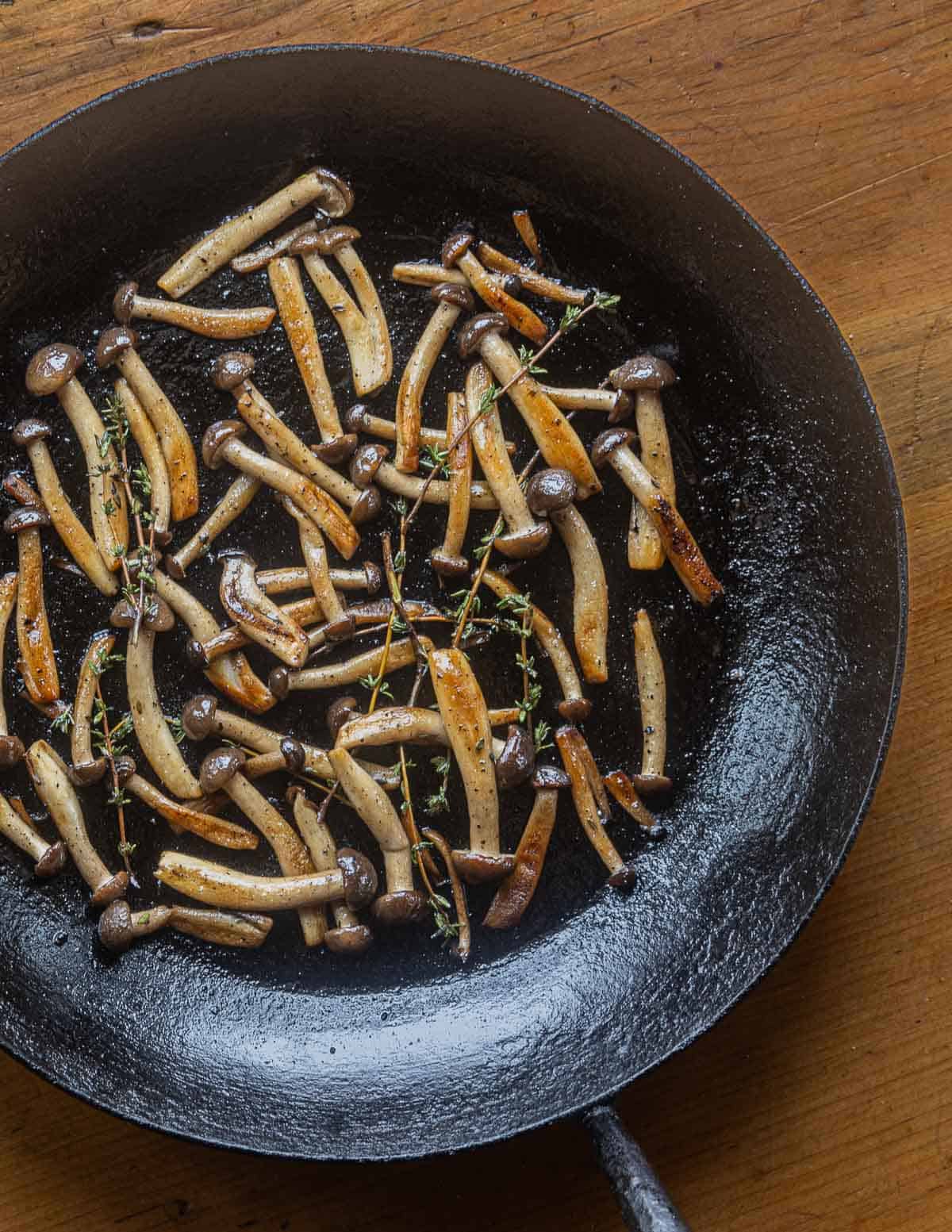 Beech mushrooms cooking with thyme sprigs in a carbon steel pan. 