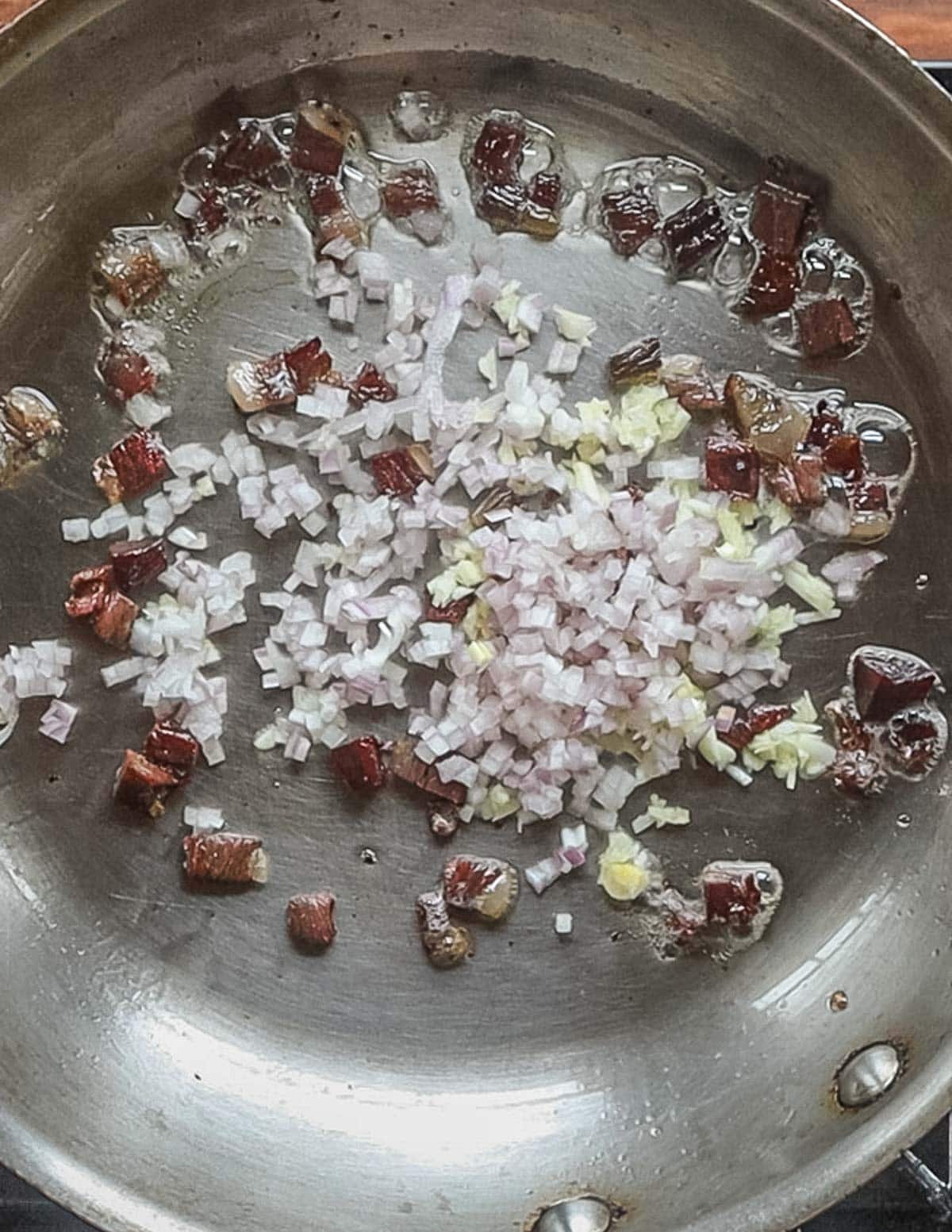 Adding diced shallots and garlic to a pan of cooking pancetta. 