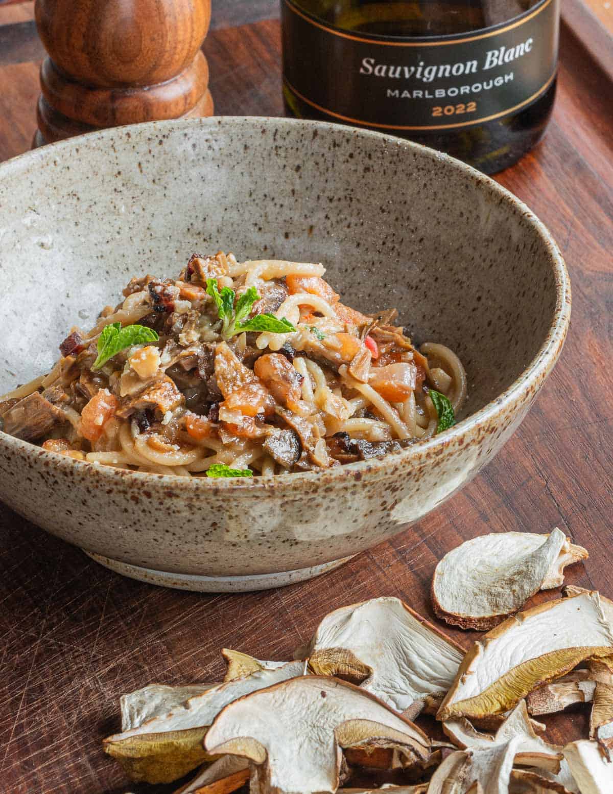 A finished bowl of porcini mushroom ragu with spaghetti in a bowl next to a bottle of wine and dried porcini mushrooms. 
