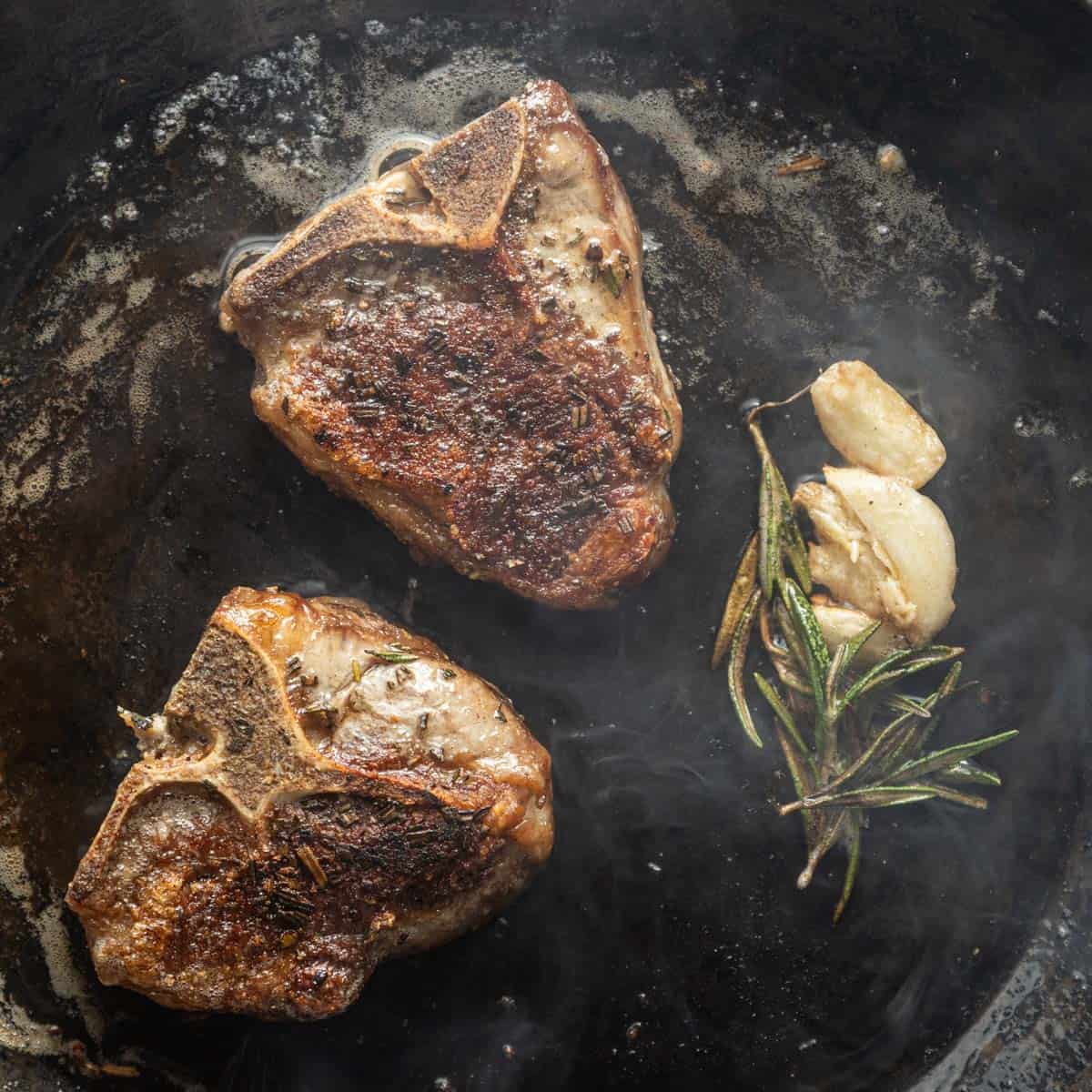 Browned goat chops cooking in a pan with garlic and rosemary. 